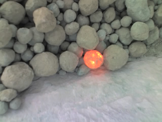 At the heart of a cluster of grey cement clinkers, one clinker glows with an orange light from within, softly illuminating its immediate vicinity. The contrast between the dull clinkers and the glowing one creates a focal point.